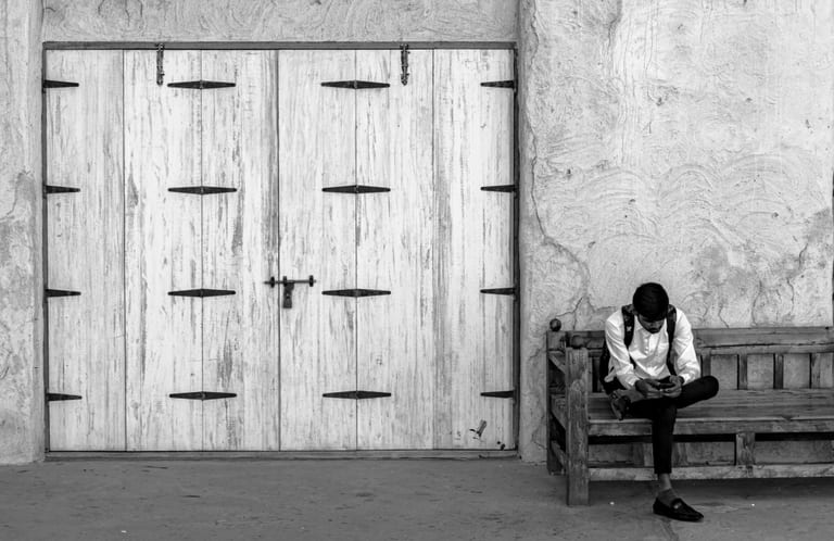 Black-and-white photograph of a person sitting on a wooden bench next to large white paneled wooden doors and a textured plaster wall, looking down at a phone.