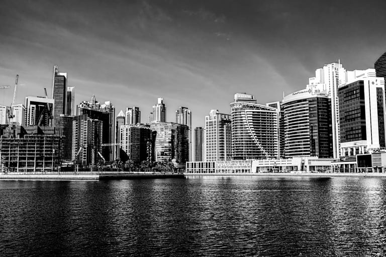 Black-and-white architectural photograph showing a dense skyline of high-rise buildings and construction cranes along a waterfront, with rippling dark water filling the foreground.