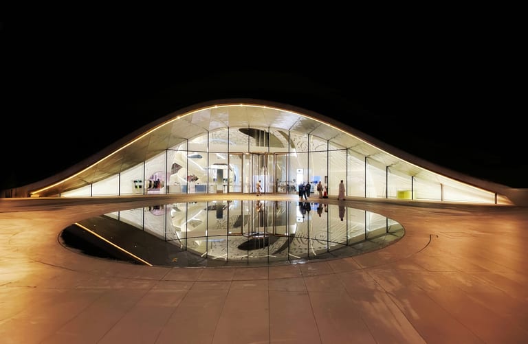 Wide night shot of a low-slung, curved glass architecture illuminated with warm interior light, featuring a perfectly circular reflecting pool in the foreground plaza and several small figures walking.