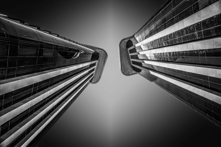 Low-angle monochrome photograph looking straight up between two tall modern buildings with gridded glass facades, creating a V-shaped framing against a dark sky.