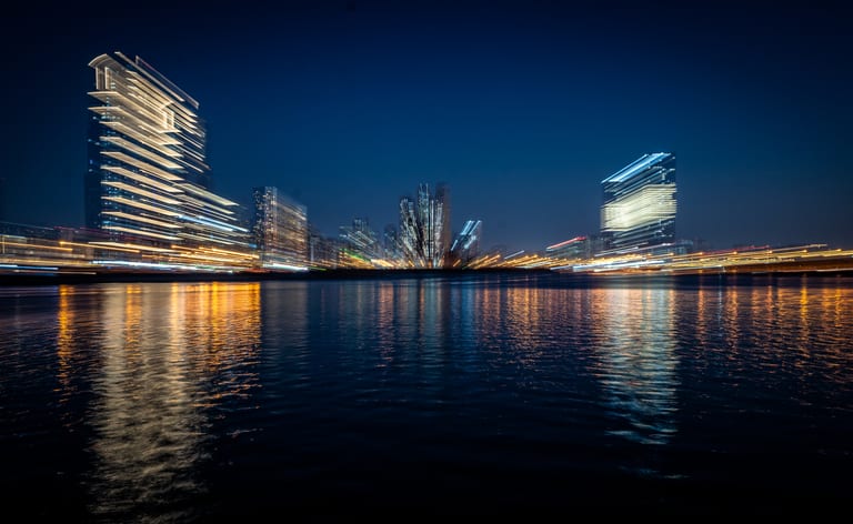 Long-exposure night photograph of illuminated skyscrapers across a body of water, featuring horizontal light streaks and blurred reflections on the water surface.