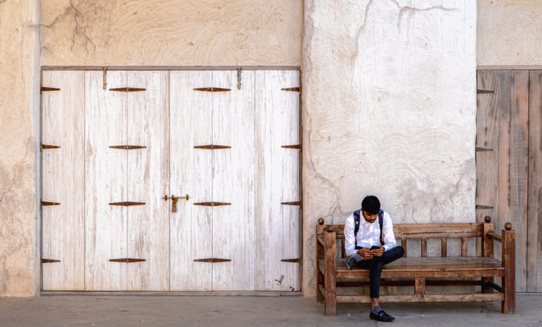 A man in a white shirt and dark trousers sits on a wooden bench against a textured plaster wall, looking down at his mobile phone next to a set of large, white-painted wooden doors.