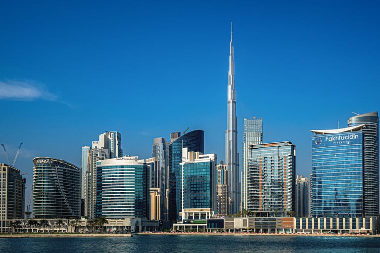 Daytime exterior shot of a modern city skyline featuring a very tall spire skyscraper rising above surrounding glass towers across a body of water.