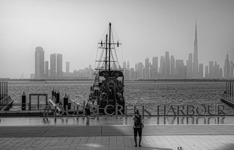 A person stands in the foreground looking at a traditional ship docked on a waterfront, with a tall city skyline in the background, captured in monochrome.
