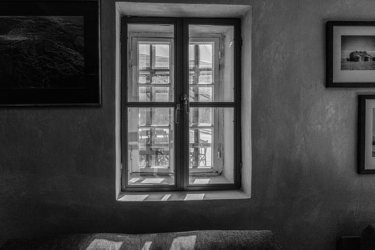 Black and white interior photograph centered on a multi-pane window in a textured wall, flanked by framed artwork, with bright light casting geometric patterns onto a sofa in the foreground.