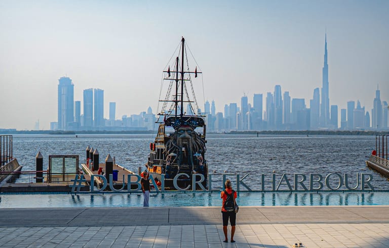 A person viewed from behind stands on a tiled waterfront plaza, facing a large, ornate ship with a carved figurehead and tall masts, with a hazy modern city skyline featuring a very tall skyscraper visible in the background across calm water.