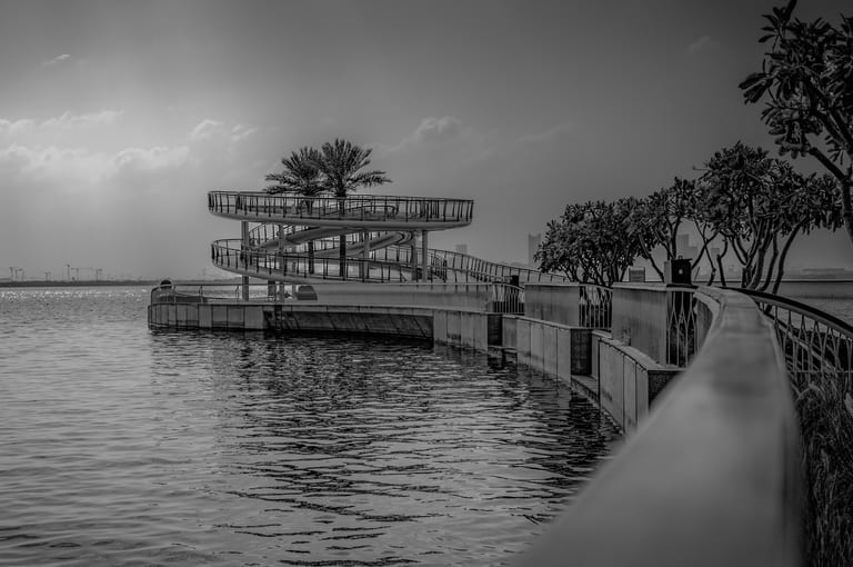 Black and white photograph of a curving spiral viewing platform over water, with palm trees on top and a paved railinged pathway leading along the shoreline.