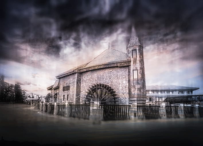 Exterior architectural view of a stone building with a large wooden water wheel and round tower under a dark, dramatic cloudy sky, with foreground motion blur.