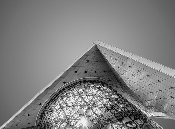 Monochrome architectural photograph shot from below, showing angular panels with circular indents and a large glass dome reflecting bright sunlight through a geometric metal lattice.