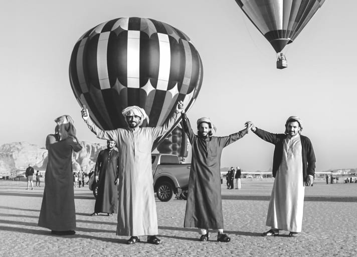Monochrome photograph of three men standing side by side holding hands in an open landscape, with two large hot air balloons floating in the bright sky behind them.