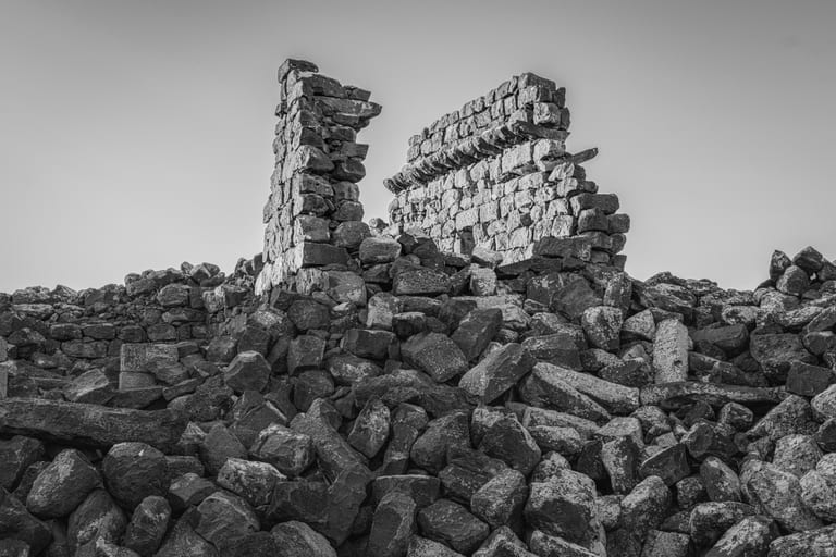 Black-and-white photograph of crumbling ancient stone walls rising behind a vast foreground pile of dark, irregular boulders under a clear sky.