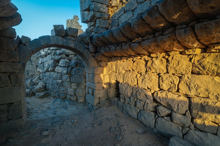 Interior view of ancient stacked stone walls and an arched entryway illuminated by golden sunset light casting strong shadows.