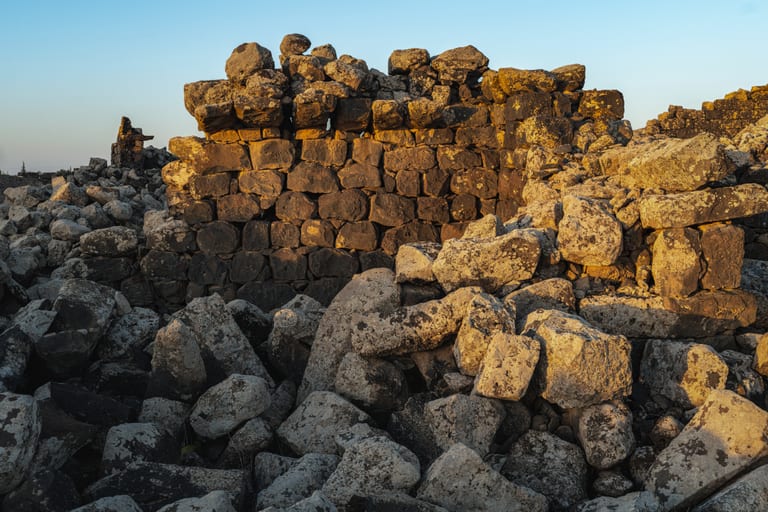An eye-level shot of ancient stacked basalt stone walls surrounded by a jumble of fallen boulders, illuminated by warm golden hour sunlight under a pale blue sky.