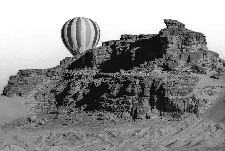 A black-and-white photograph showing a striped hot air balloon appearing just behind a massive, textured sandstone cliff face under a pale sky.