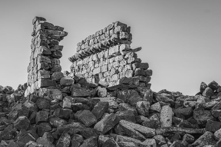 Black and white photograph of ancient stone ruins and rubble with dark basalt blocks piled in the foreground against a pale sky.