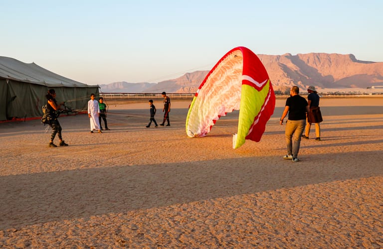 Several people stand around a large red, white, and yellow paraglider wing on a dry desert plain beside a large green tent, with rugged mountains under a clear sky in the background.