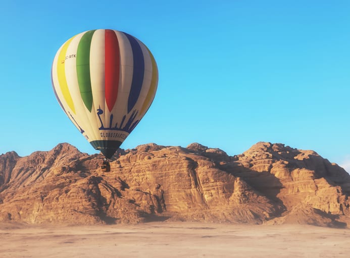 A multi-colored striped hot air balloon floats in the upper left of the frame over a low rocky mountain range and flat desert sand under a blue sky.