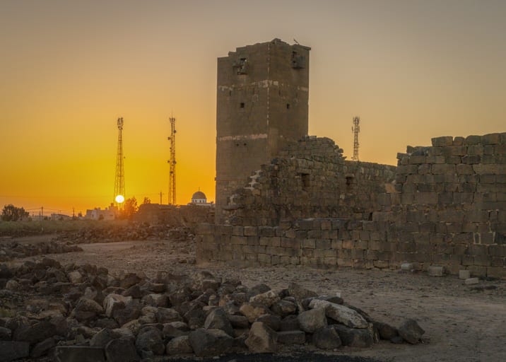Low-angle photograph of ancient stone ruins and a tall tower against a vibrant yellow sunset sky, with scattered dark rocks in the foreground and metal communication towers on the horizon.
