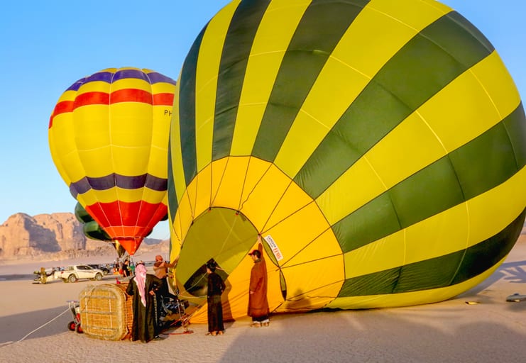 Low-angle shot of a giant green and yellow striped hot-air balloon resting on desert sand with a smaller balloon and several figures standing near the wicker basket.