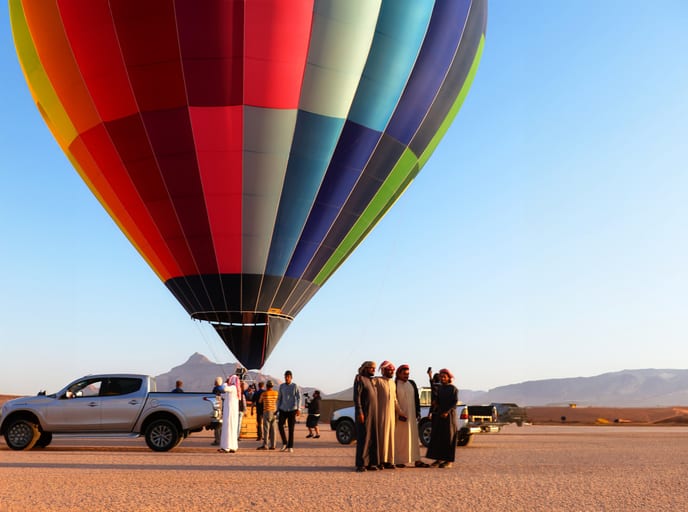 Low-angle shot of a massive, multi-colored striped hot air balloon filling the upper half of the frame above people standing near pickup trucks on a flat, arid landscape.