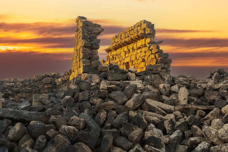 An ancient stone wall ruin standing over a field of dark volcanic rocks under a golden sunset sky.