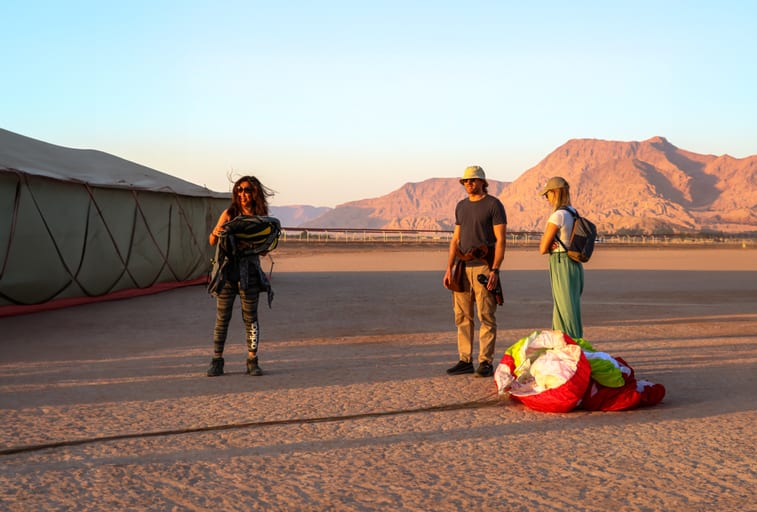 Three people stand outdoors on flat desert ground in low-angle sunlight. On the left, a person holds a piece of gear near a large tent-like structure. In the center and right, two people stand near a bundled red, white, and green paraglider. Rugged mountains are visible in the background.