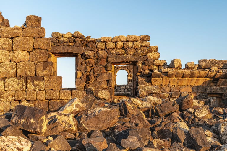 A weathered stone wall featuring two window openings with rubble scattered across the foreground, captured in warm sunset light under a clear sky.