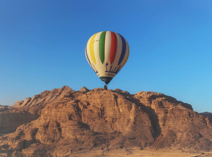 A large multi-colored hot air balloon hovers in a clear blue sky just above a jagged, sunlit rocky mountain range.