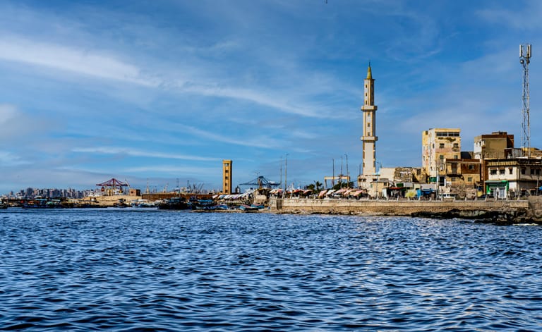 Wide exterior view of a coastal cityscape featuring a prominent tall stone minaret next to older buildings along a seawall, with choppy dark blue water in the foreground and a bright blue sky overhead.