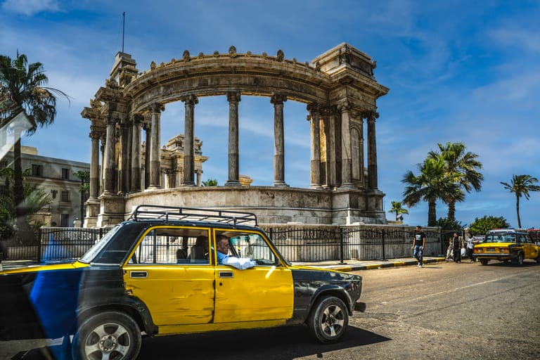 Street level view featuring a yellow and black taxi cab driving across a paved road, passing an ornate semicircular stone monument with towering classical columns and palm trees under a bright blue sky.