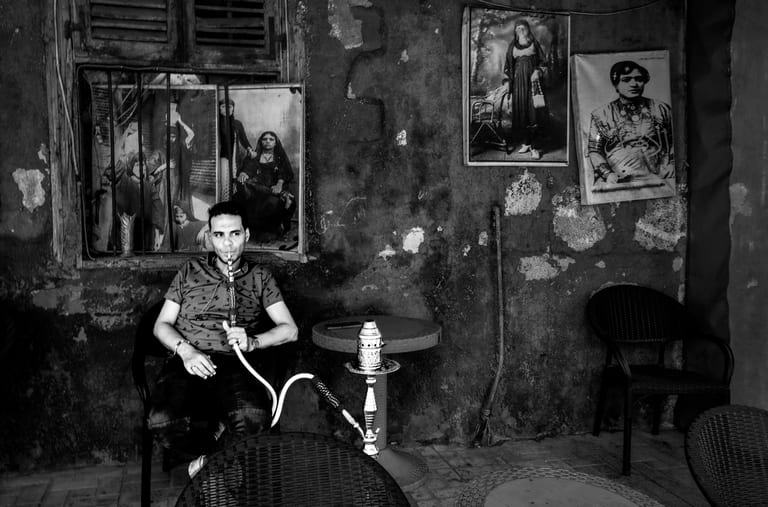 A black-and-white street photograph of a man seated in a dark chair, smoking a shisha pipe. He is surrounded by weathered, textured walls adorned with framed vintage portraits, with an empty table and chairs nearby.