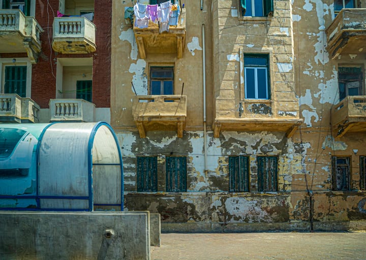 Exterior architectural view of a weathered multi-story building facade with peeling plaster, wooden shutters, small balconies, and hanging laundry, alongside a curved blue structure in the lower left foreground.