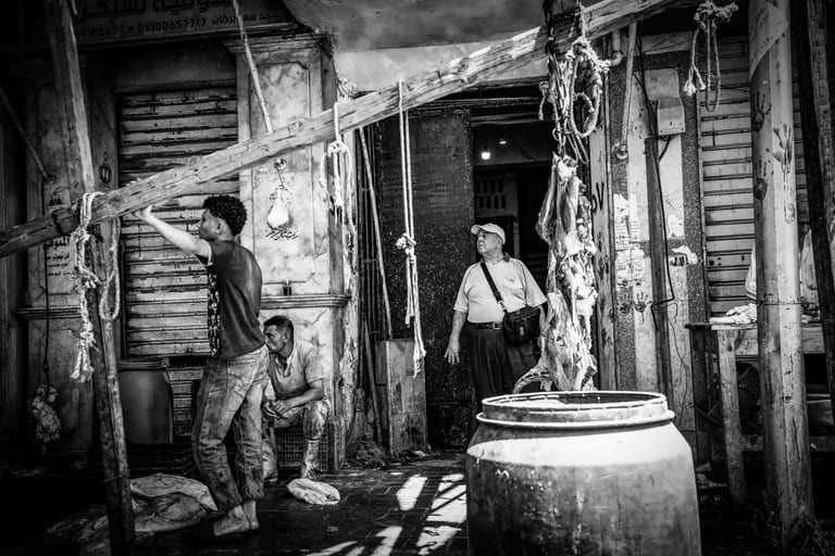 Black and white street photograph of an outdoor market scene in Alexandria, showing a young worker holding a wooden beam on the left, a seated man behind him, and an older man standing in a doorway near hanging ropes and a large plastic drum.