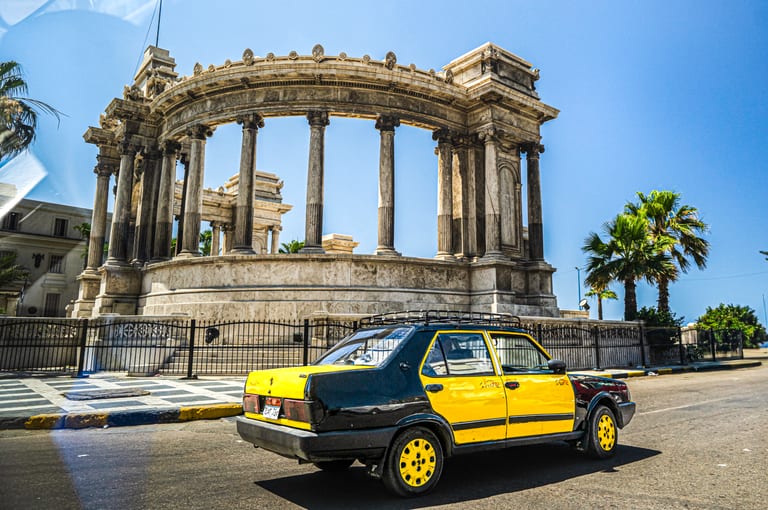 A yellow and black taxi parked on an asphalt street in front of a grand, curved neoclassical stone colonnade structure under a clear blue sky, with palm trees visible on the right.