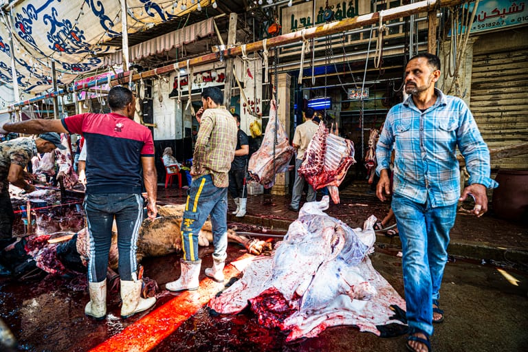 A busy market street scene with several men working amongst hanging meat carcasses and large animal hides spread across a wet red-stained floor.