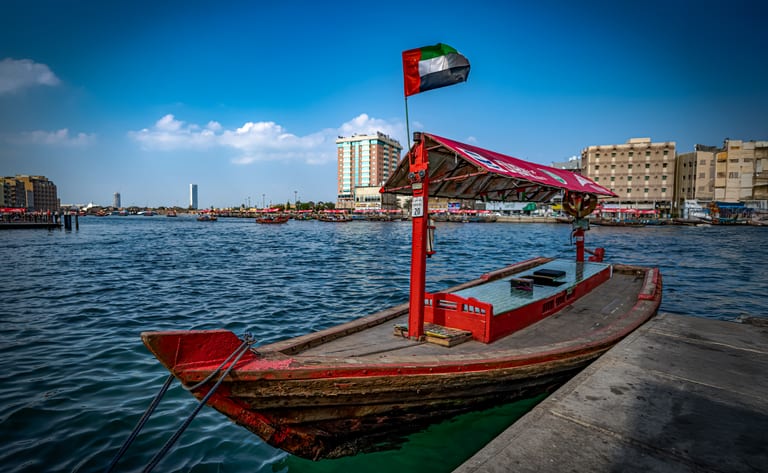 A traditional wooden water taxi with a red canopy and a national flag is tied to a concrete dock, floating on deep blue water with buildings visible across the waterway.