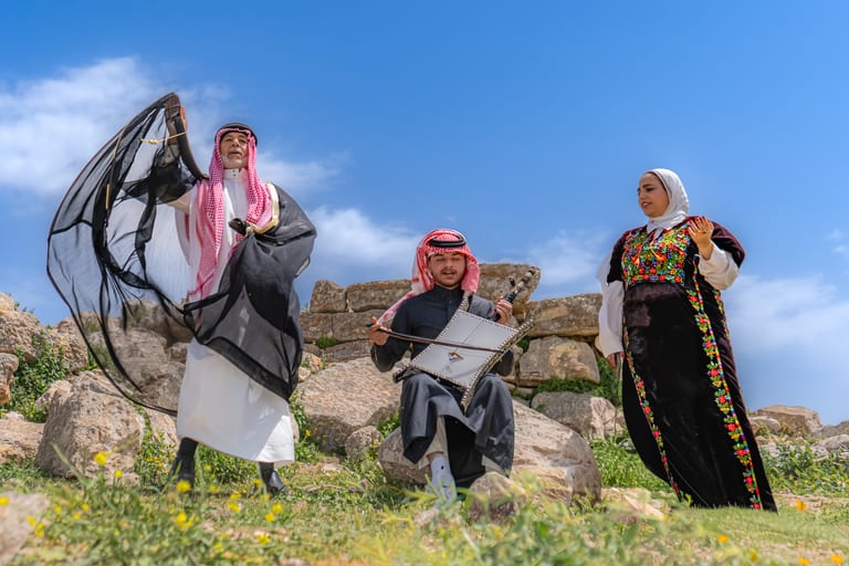 A low-angle outdoor shot of three people in traditional dress: a man standing on the left waving a dark sheer cloth, a seated young man playing a square string instrument in the center, and a woman standing on the right in an embroidered gown.
