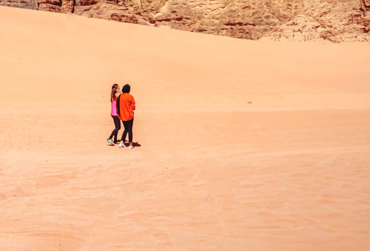 Two people wearing bright orange and pink jackets walking together across a vast orange sand dune with rocky cliffs visible in the background.