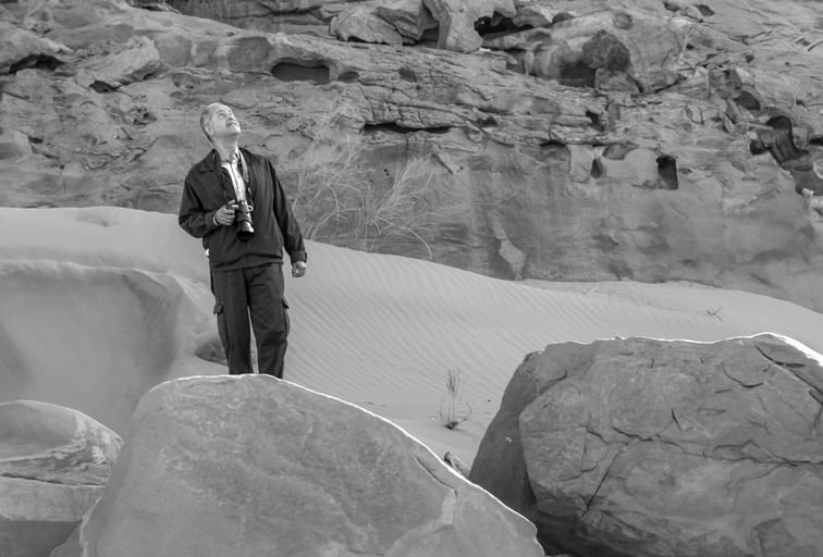 Monochrome photograph of a man holding a camera, standing among large desert rocks and sand dunes while looking upward.