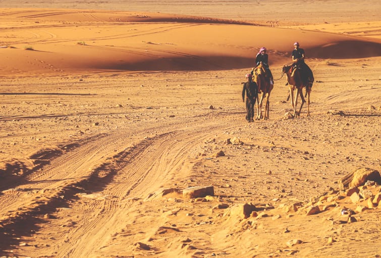 Wide-angle outdoor shot of two people riding camels and one person walking across a vast sand and rock desert landscape under warm sunlight.