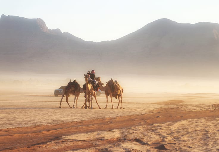 Wide-angle landscape view of three saddled camels with riders standing together in a sandy desert plain, surrounded by a heavy morning haze and large mountains under a bright sky.