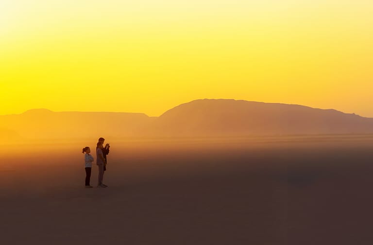 Three people standing in profile on a vast desert floor against a bright yellow sunset sky, with layered mountain silhouettes in the background.