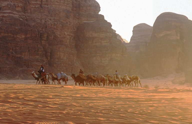 A line of multiple camels carrying riders and baggage walks across a desert sand dune landscape, framed by massive rocky cliffs under a bright, hazy sky.