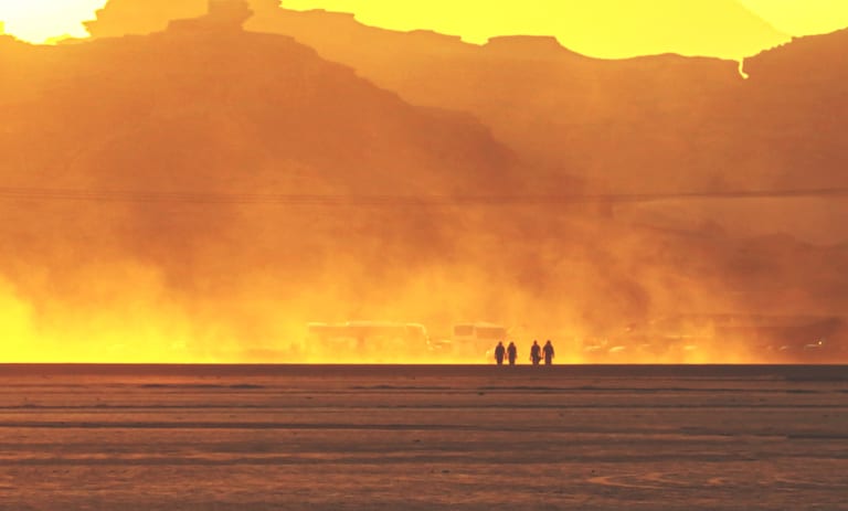 Four silhouetted figures walking across a flat ground amidst a heavy cloud of golden dust and bright backlit sky, with dark mountainous terrain rising in the upper background.