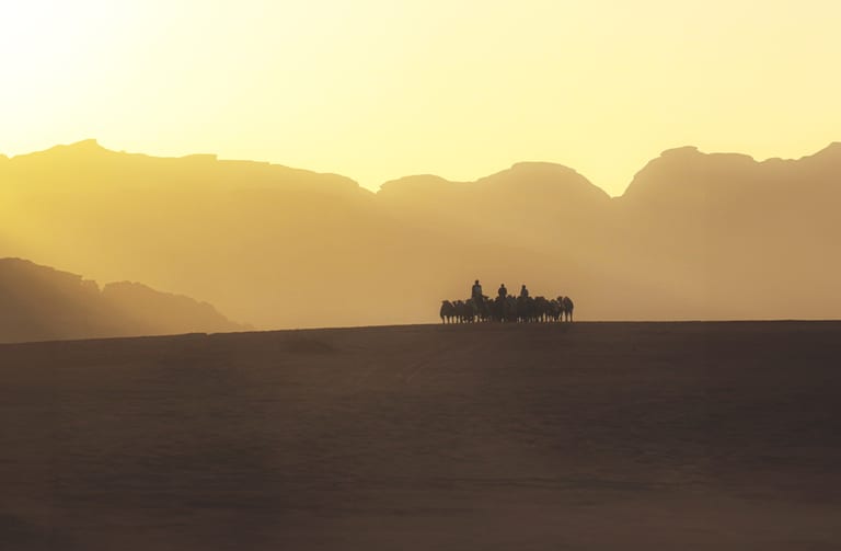 Silhouette of a caravan of people riding camels crossing a dark desert ridge in front of a hazy golden sunset and distant mountain silhouettes.