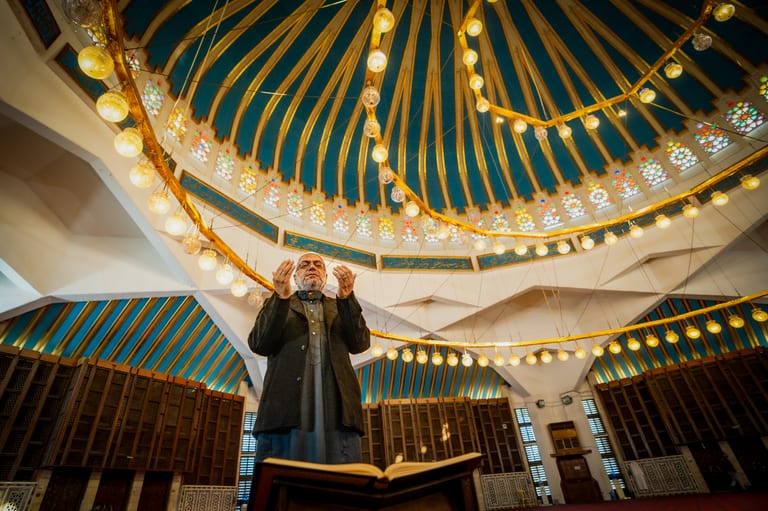 An elderly man standing inside a large hall with arms raised in prayer beneath a high domed ceiling featuring radial ribs and circular illuminated chandeliers.