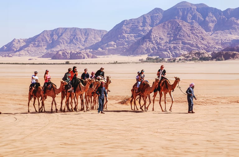A group of people riding camels in a wide desert landscape with rocky mountains in the background, led by guides walking on the sand.